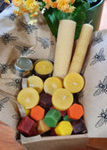 Assorted small candles in a box with beeswax candles on top, on a tablecloth with bee pattern.