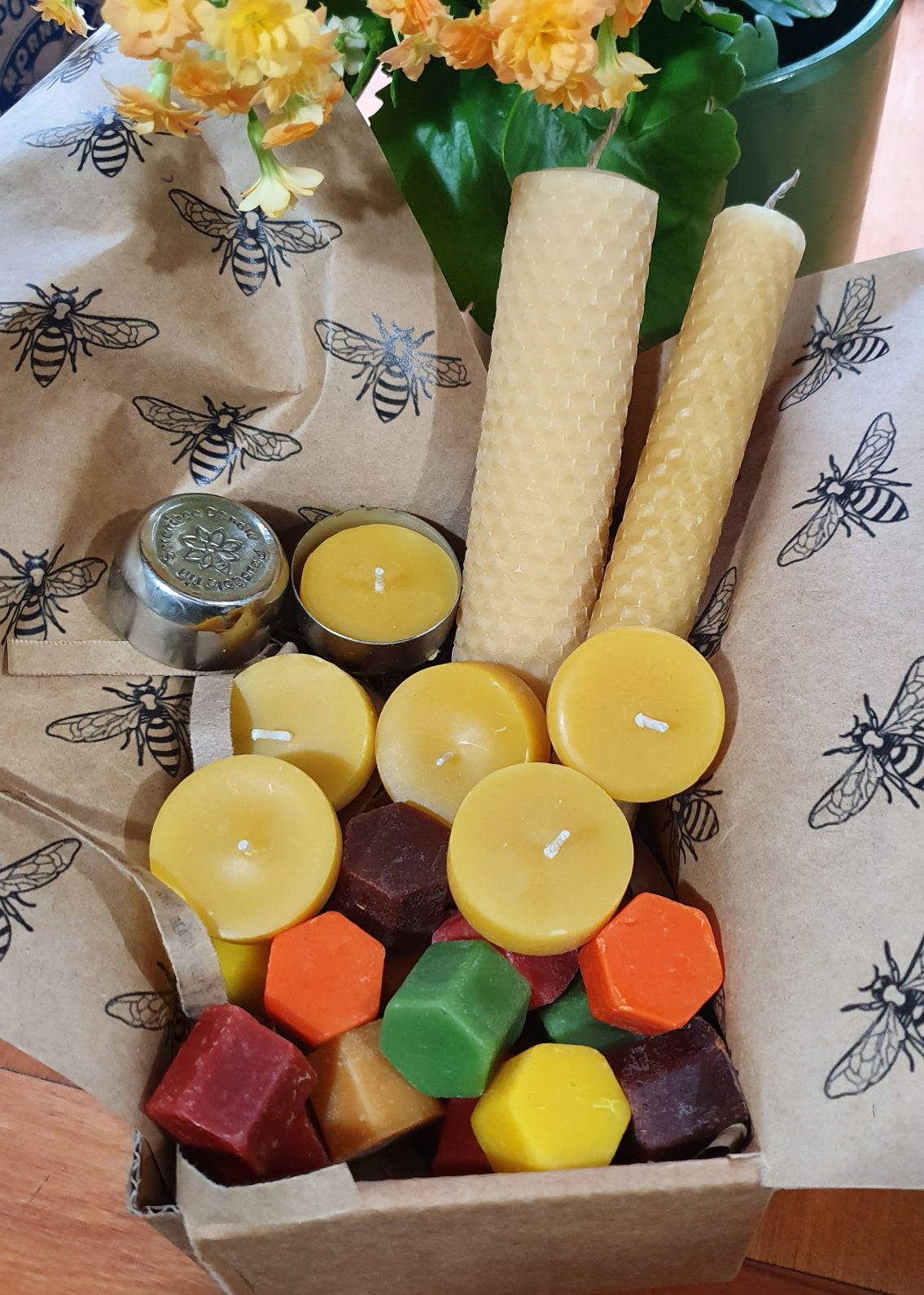 Assorted small candles in a box with beeswax candles on top, on a tablecloth with bee pattern.