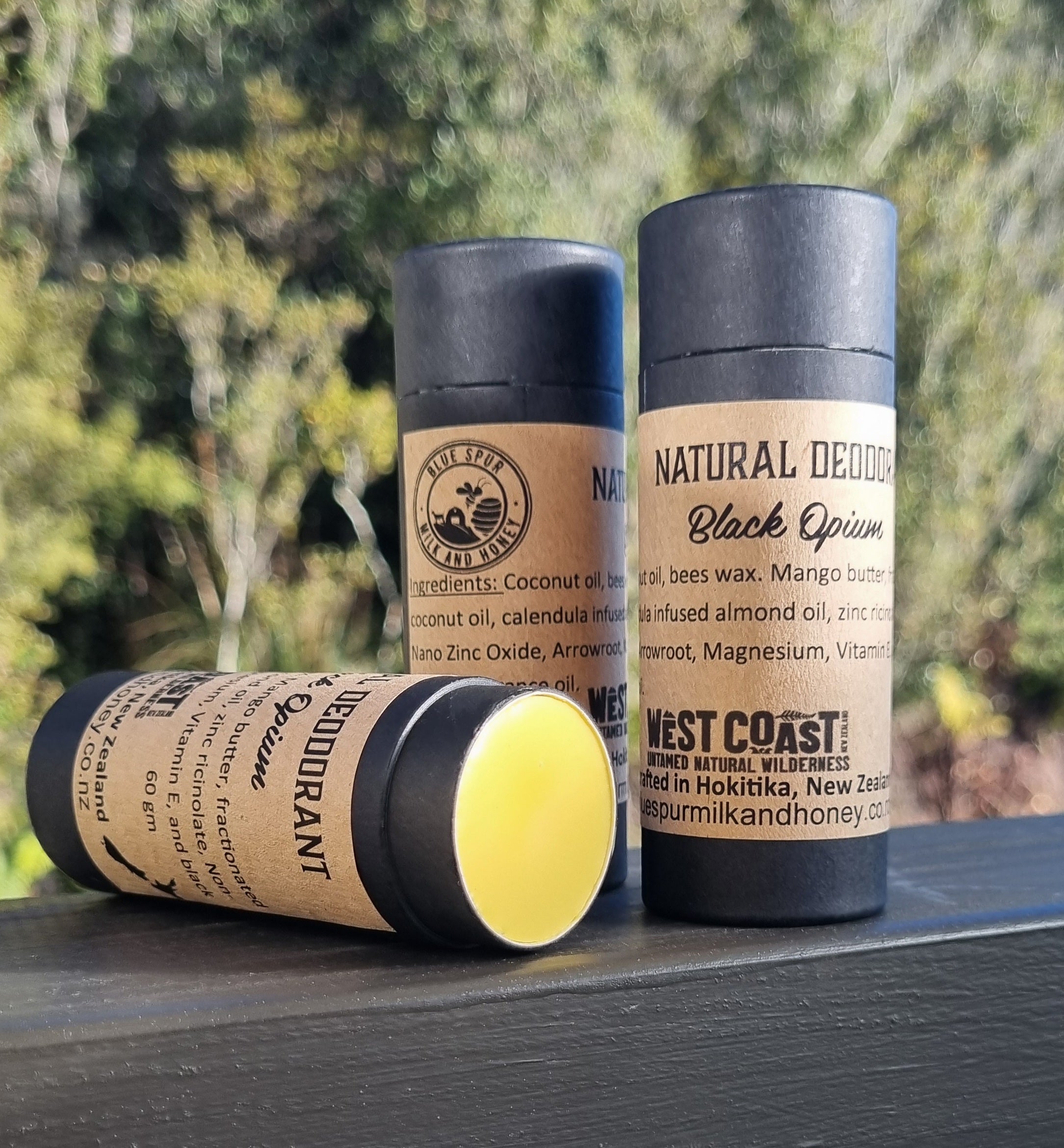 Three natural deodorant sticks with black caps and cardboard labels on a wooden surface with a blurred green background.