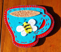 Decorative hand stitched felt coaster with a blue cup with a red rim and handle, featuring a bee design on a wooden surface.