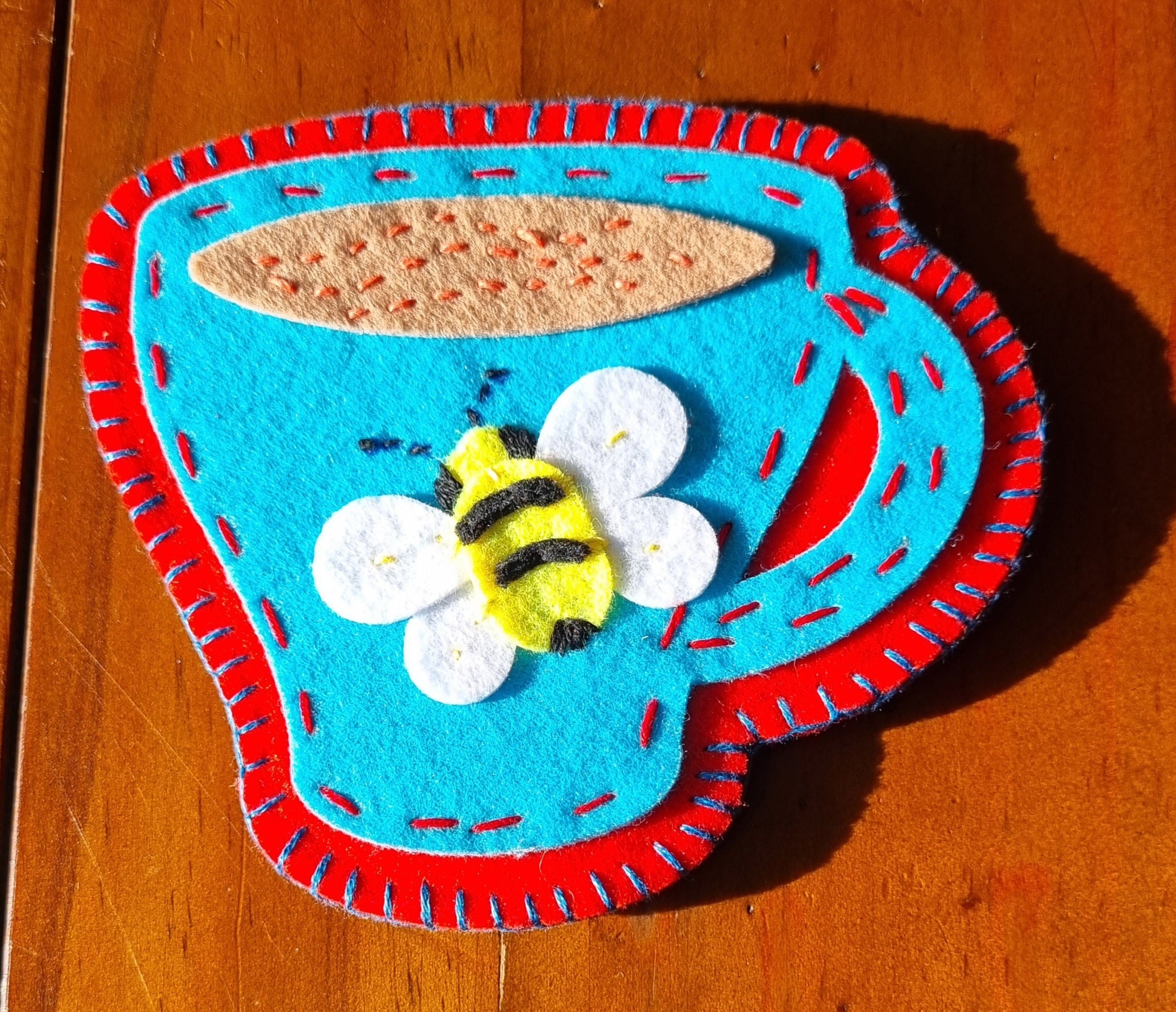Decorative hand stitched felt coaster with a blue cup with a red rim and handle, featuring a bee design on a wooden surface.