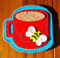 Decorative hand stitched felt coaster with a red cup with a blue rim and handle, featuring a bee design on a wooden surface.