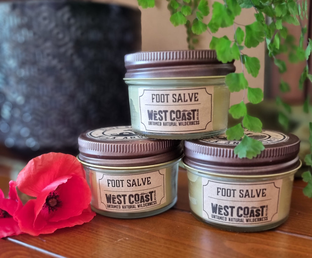 Three jars of West Coast Foot Salve on a wooden surface with a pink flower and green leaves in the background.