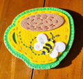 Decorative hand stitched felt coaster with a yellow cup with a green rim and handle, featuring a bee design on a wooden surface.