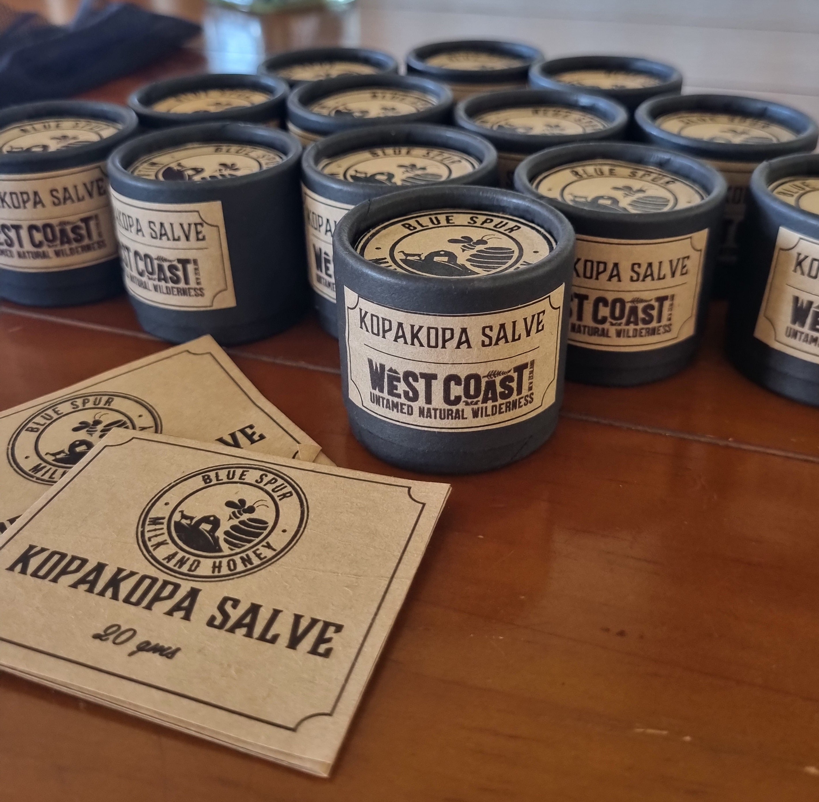 Multiple containers of West Coast Kopakopa Salve on a wooden surface with branded cards.