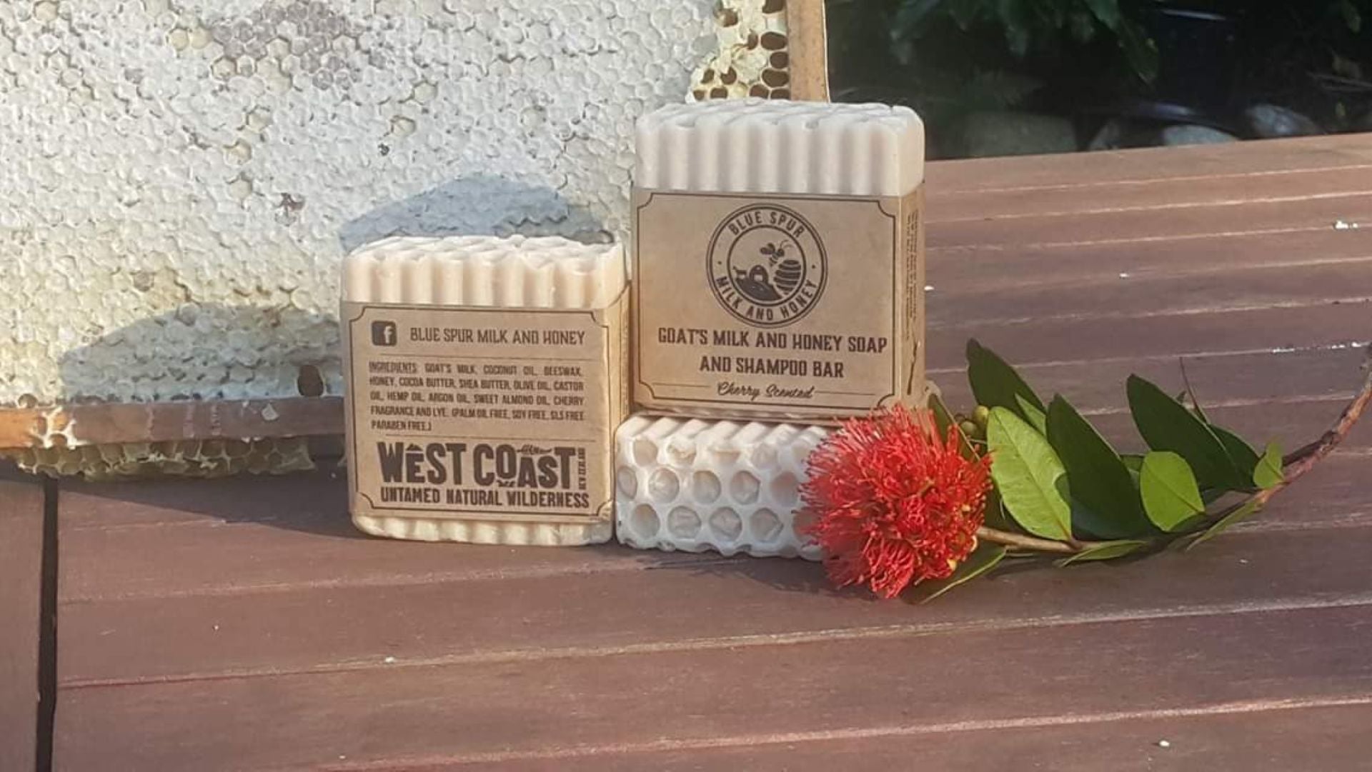 Two soap bars with 'West Coast' branding on a wooden surface with a red flower and green leaves.