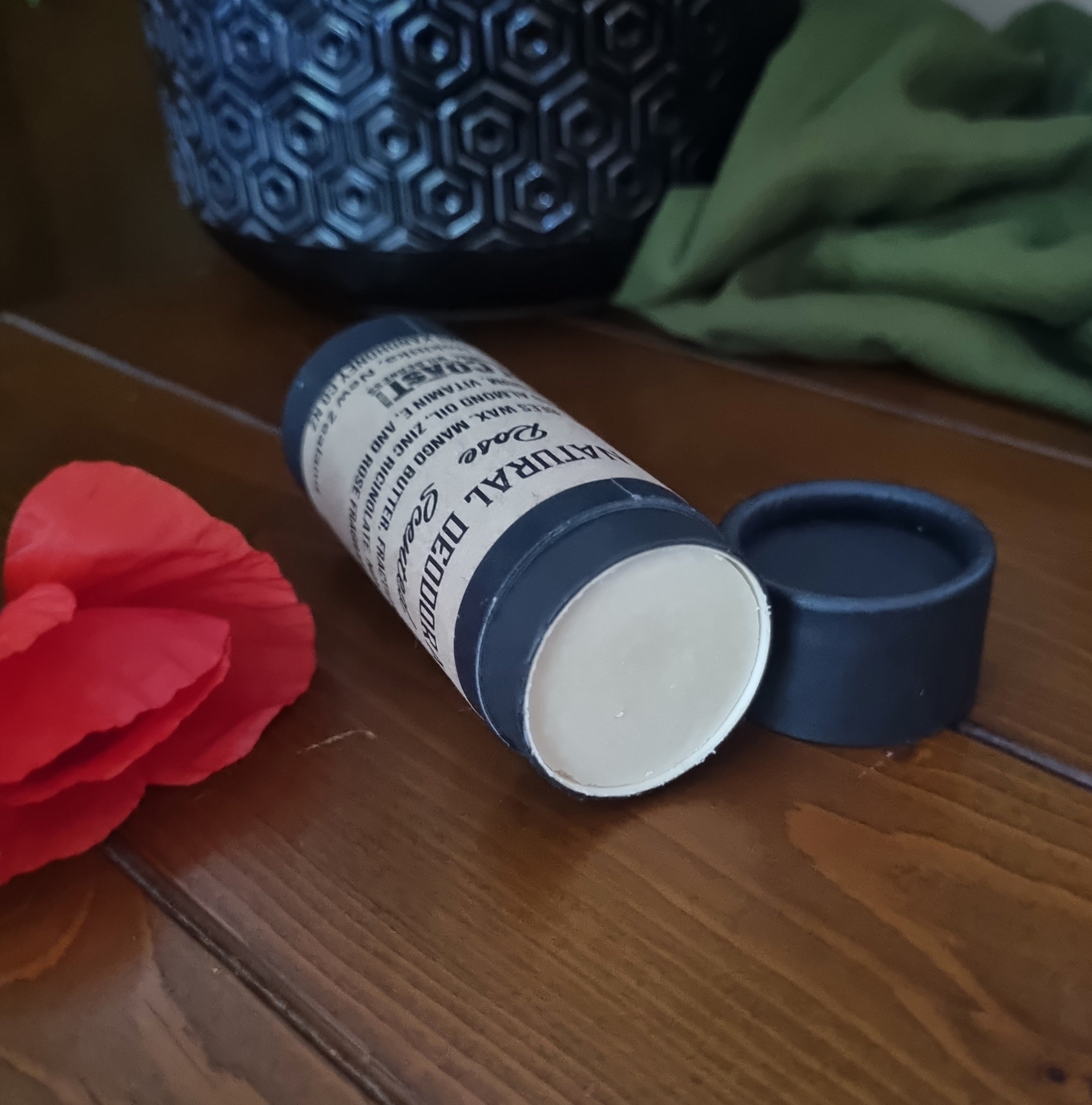 
open rose deodorant in black paperboard tube lying on its side with cap off laying on wooden table with a red poppy