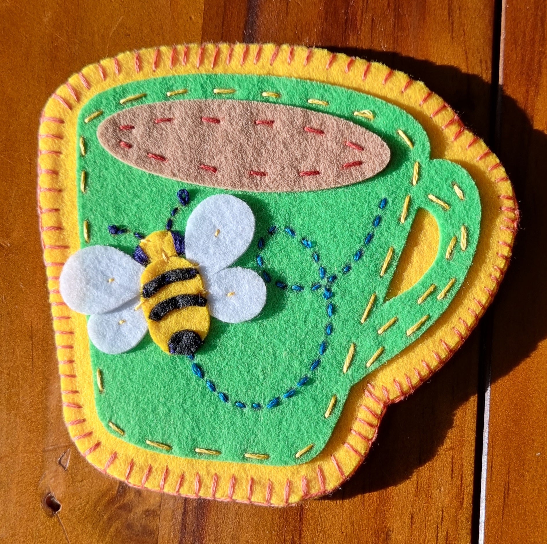 Decorative hand stitched felt coaster with a green cup with a yellow rim and handle, featuring a bee design on a wooden surface.